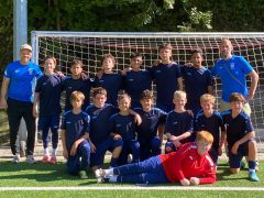 Die U16-Fußballmannschaft der Adolf-Reichwein-Schule steht gemeinsam mit ihren beiden Trainern vor einem Fußballtor. Die Spieler tragen dunkelblaue Trikots und Shorts. Im Vordergrund liegt der Torwart in einem roten Trikot auf dem Kunstrasen. Links steht Sportlehrkraft und Trainer Herr Strasser, rechts Sportlehrkraft und Trainer Herr Schulz. Die Mannschaft und ihre Trainer schauen in die Kamera. Im Hintergrund sind das Fußballtor und Bäume zu sehen.