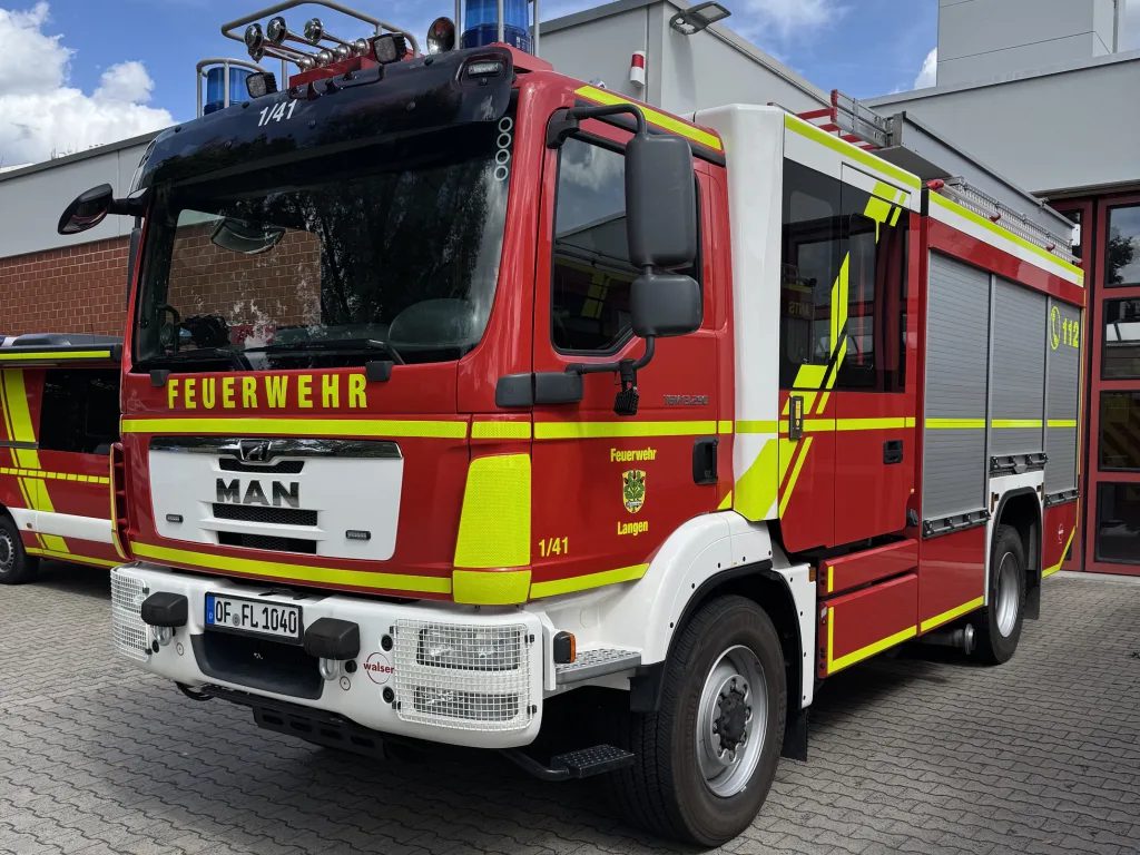 Ein rotes Löschfahrzeug der Feuerwehr Langen steht auf dem Hof der Feuerwache. Das Einsatzfahrzeug ist von vorne und von der rechten Seite zu sehen. Auf der Fahrzeugfront steht „FEUERWEHR“, an der Seite sind das Wappen und der Schriftzug „Feuerwehr Langen“ angebracht. Das Fahrzeug verfügt über eine große Mannschaftskabine, seitliche Geräteräume mit geschlossenen Rollläden und eine auffällige gelb-grüne reflektierende Beklebung. Im Hintergrund sind Teile der Feuerwache und ein blauer Himmel mit Wolken zu sehen.