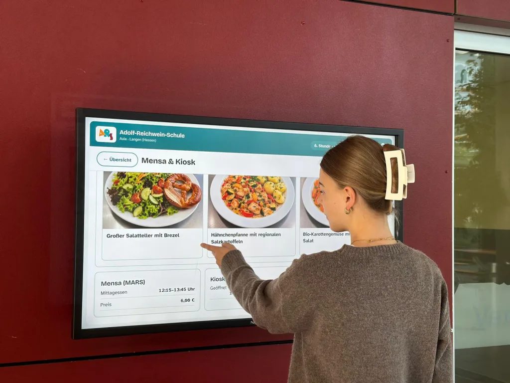 Fotografische Aufnahme einer Schülerin bei der Bedienung des neuen Touchscreen-Displays in der Aula der Adolf-Schule. Links oben auf dem Bildschirm ist das Logo der Adolf-Reichwein-Schule zu sehen. Auf dem großen Display steht in weißen und dunklen Buchstaben die Benutzeroberfläche „Mensa & Kiosk“. In der Mitte tippt eine Schülerin mit dem Finger auf das Auswahlfeld „Großer Salatteller mit Brezel“, zu dem eine entsprechende Abbildung angezeigt wird. Rechts daneben sieht man weitere Gerichte der Mensa. Die Schülerin trägt einen braunen Pulli, Goldschmuck und eine helle Haarklammer. Im unteren Bereich des Touchscreens sind Details zum Menü wie die Mensa-Öffnungszeiten und der Preis aufgelistet.
