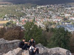 Von einer alten Bruchsteinmauer aus geht der Blick über bewaldete Hänge auf einen Ort mit vielen Wohnhäusern. Drei Schüler sitzen auf der Mauerkrone und schauen ins Tal.
