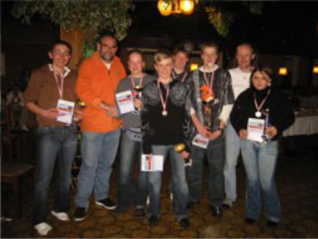 Die Gewinner des Skirennens stehen mit Medaillen und Urkunden zwischen zwei erwachsenen Organisatoren in einem Innenraum.