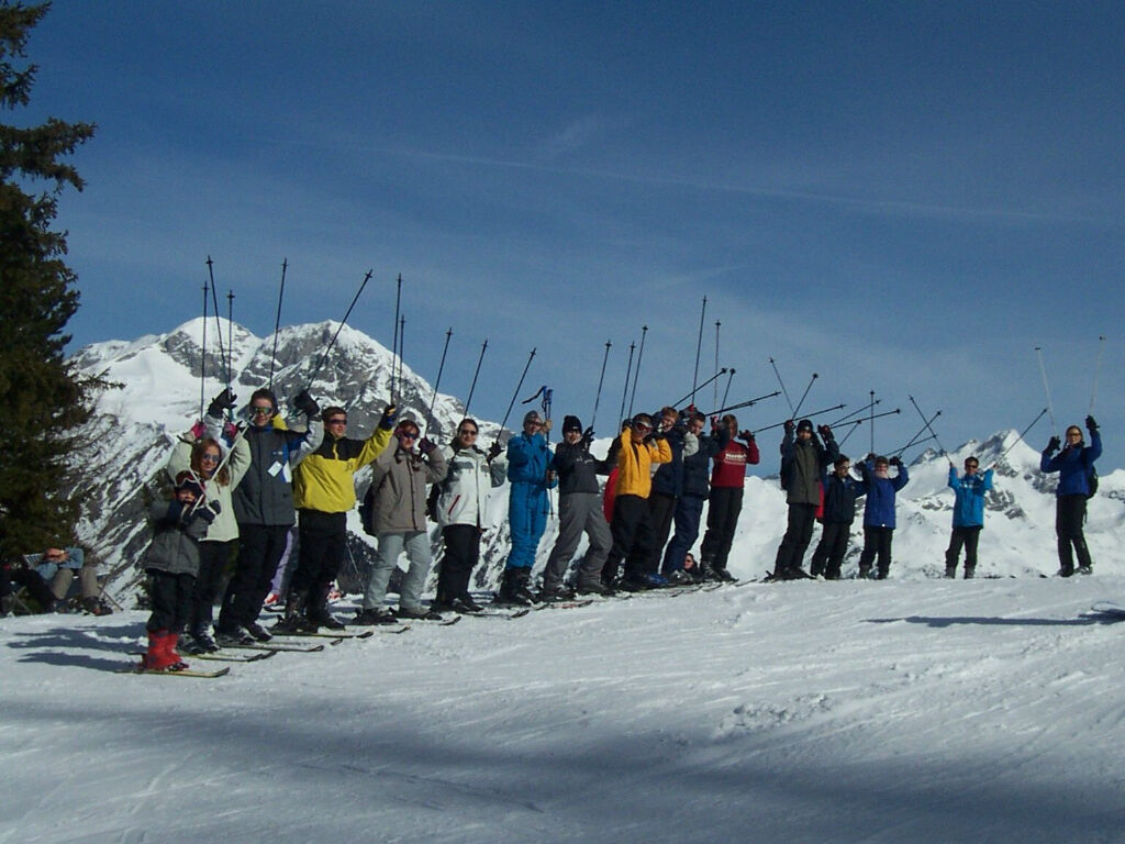 Eine Gruppe auf Skiern steht in einer Reihe auf einer Piste und reckt jubelnd die Skistöcke in die Höhe; im Hintergrund schneebedeckte Alpengipfel unter blauem Himmel.