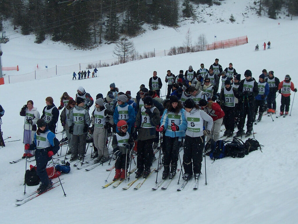 Eine große Gruppe auf Skiern mit Startnummern steht bereit auf einer verschneiten Piste; im Hintergrund ein Skihang mit Wald.