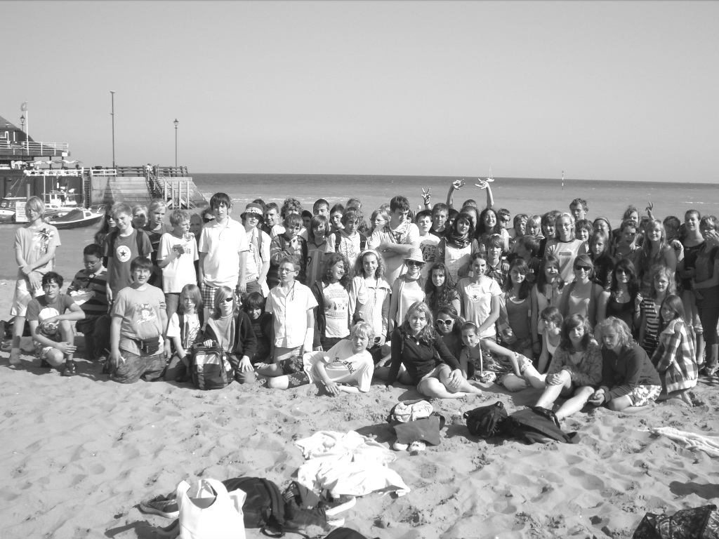 Eine große Schülergruppe posiert an einem Sandstrand; im Hintergrund ein Anleger und das Meer (Schwarz-Weiß-Aufnahme).