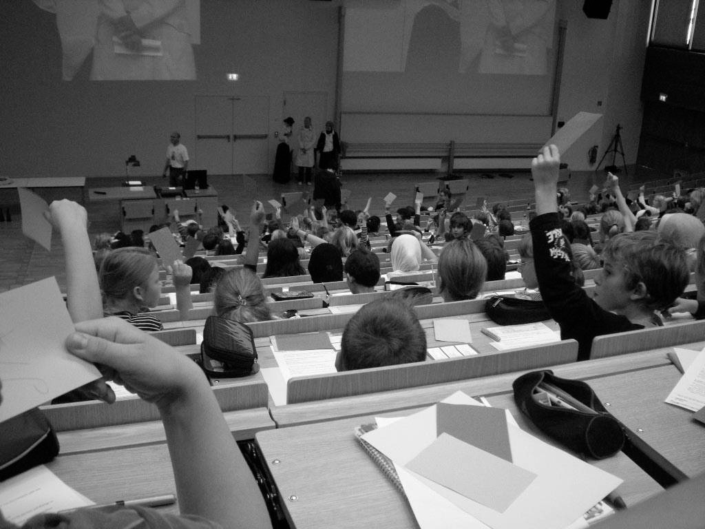 Kinder sitzen in einem großen Universitäts-Hörsaal und halten Antwortkarten hoch; vorne stehen die Vortragenden (Schwarz-Weiß-Aufnahme).