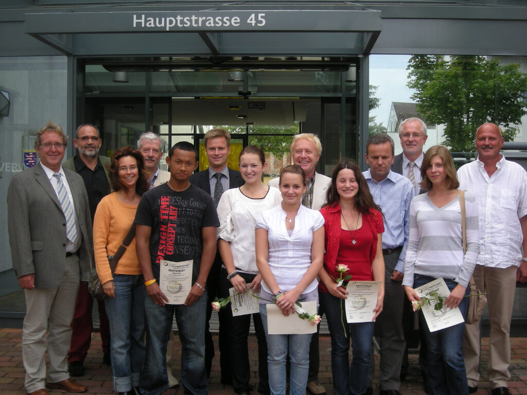 Mehrere junge Menschen halten Zertifikate und Rosen in den Händen, umgeben von mehreren Erwachsenen, vor dem Eingang eines Gebäudes mit der Aufschrift „Hauptstrasse 45“.