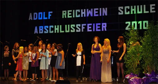 Auf der Bühne steht die Abschlussgruppe vor dem beleuchteten Schriftzug „Adolf-Reichwein-Schule Abschlussfeier 2010“.
