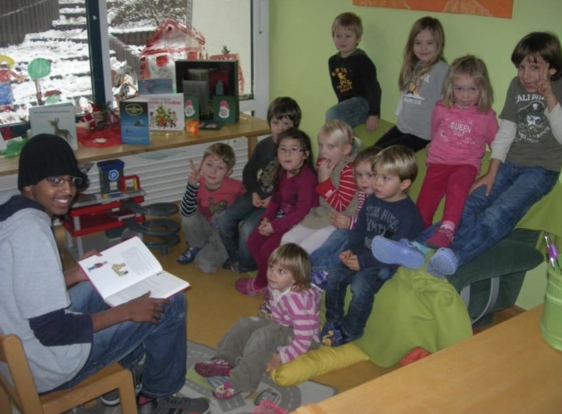 Eine jugendliche Person liest einer Gruppe kleiner Kindergartenkinder aus einem Bilderbuch vor; auf einer Ablage stehen ein Lebkuchenhaus und weihnachtliche Deko.