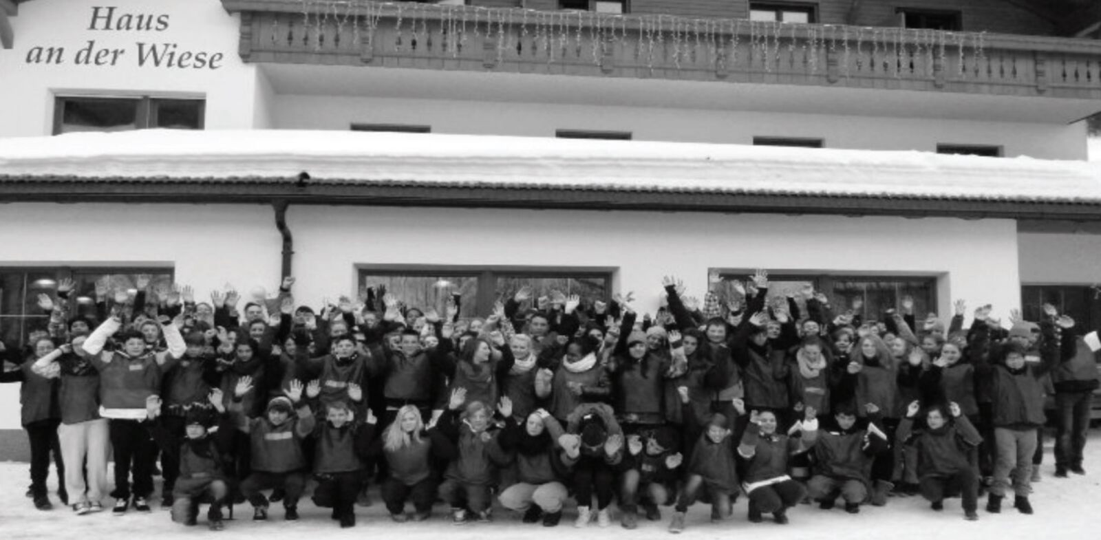 Eine sehr große Gruppe in Winterkleidung winkt vor einem verschneiten Alpen-Gästehaus mit der Aufschrift „Haus an der Wiese“ (Schwarz-Weiß-Aufnahme).
