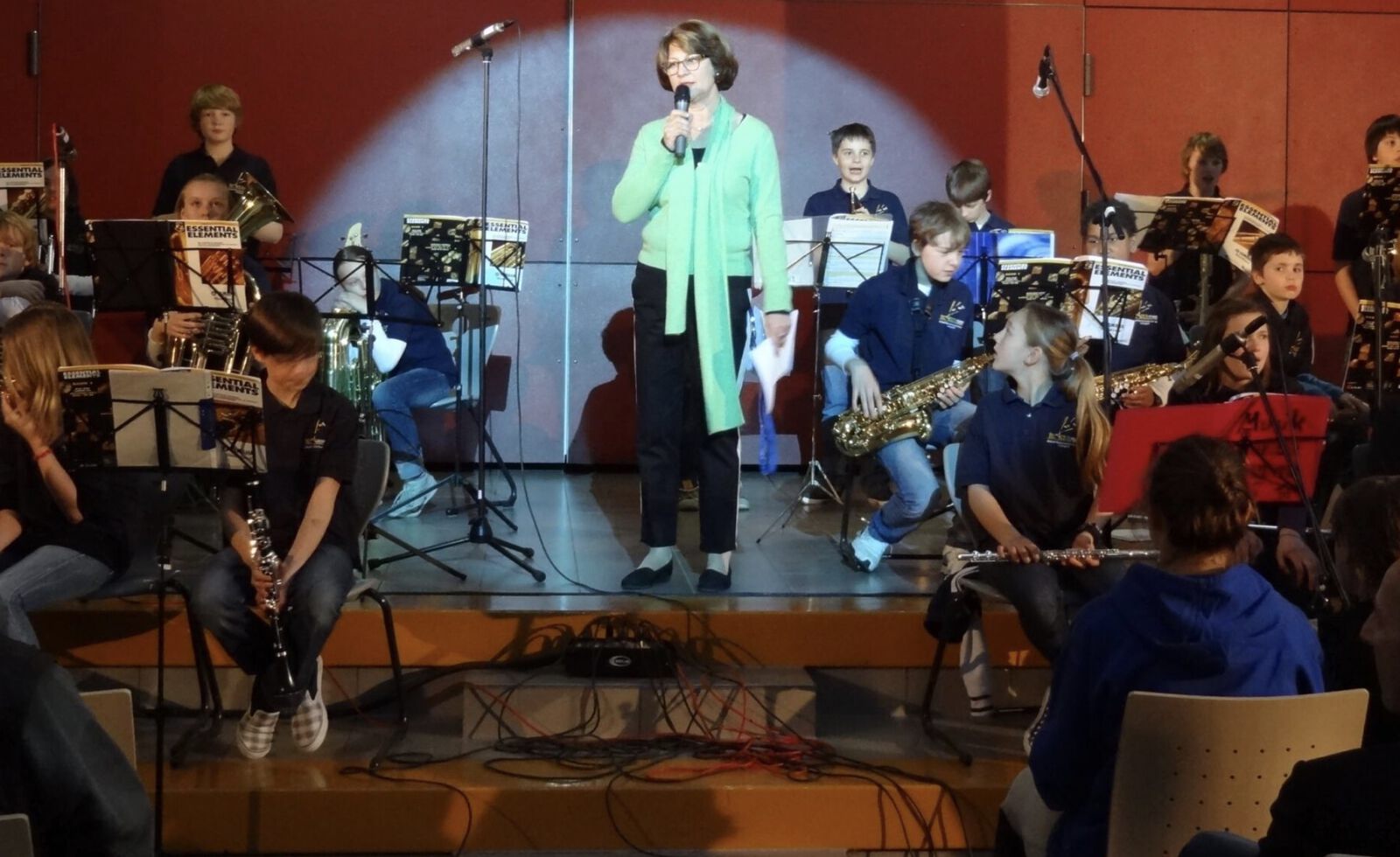 Eine Frau steht im Scheinwerferlicht auf der Bühne und spricht ins Mikrofon; um sie herum sitzt eine Schul-Big-Band mit Blasinstrumenten wie Saxophonen, Tuba und Klarinetten an Notenständern.