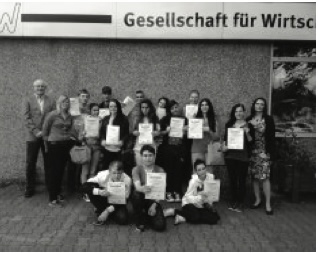 Eine Schulklasse hält Urkunden in die Kamera, an den Seiten zwei Erwachsene, vor einem Gebäude mit der Aufschrift „Gesellschaft für Wirtschaft…“ (Schwarz-Weiß-Aufnahme).