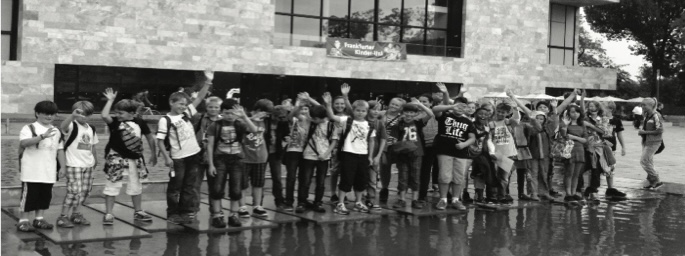 Eine große Gruppe Kinder winkt vor einem Universitätsgebäude mit dem Banner „Frankfurter Kinder-Uni“; davor ein flaches Wasserbecken (Schwarz-Weiß-Aufnahme).
