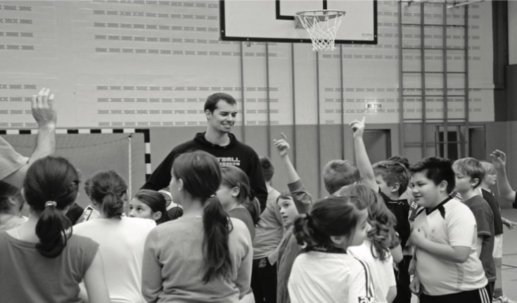 Ein großgewachsener Mann steht in einer Sporthalle umringt von vielen Kindern; einige strecken die Hand hoch, über ihnen ein Basketballkorb (Schwarz-Weiß-Aufnahme).