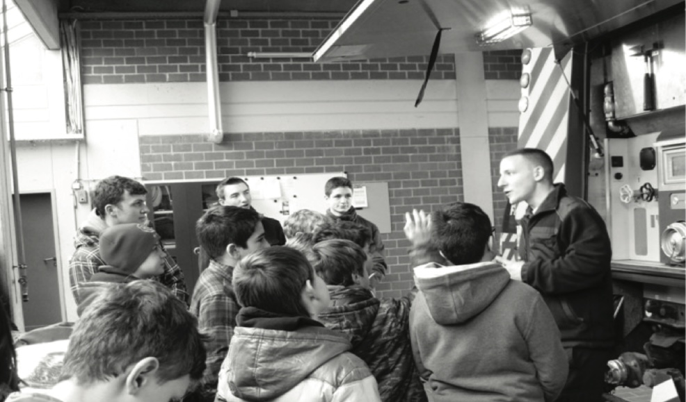 Eine Gruppe Kinder steht in einer Feuerwache um eine erwachsene Person, die neben dem Bedienfeld eines Feuerwehrfahrzeugs etwas erklärt (Schwarz-Weiß-Aufnahme).