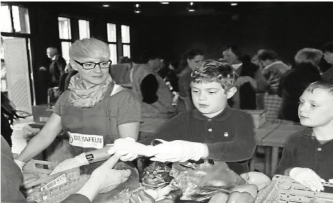 Zwei Jungen reichen gemeinsam mit einer erwachsenen Person in Tafel-Schürze Lebensmittel über einen Ausgabetisch; im Hintergrund weitere Menschen (Schwarz-Weiß-Aufnahme).