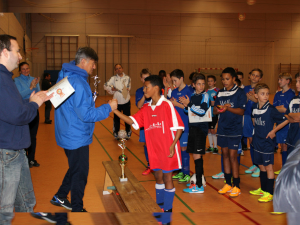 Auf dem Bild ist ein Mann in blauer Jacke zu sehen, der einem jungen Schüler mit einem Pokal gratulieren will. Im Hintergrund stehen andere Jungen im Fußballtrikot. Die Szene wirkt sehr positiv und die Freude des jungen Schüler ist deutlich zu erkennen.