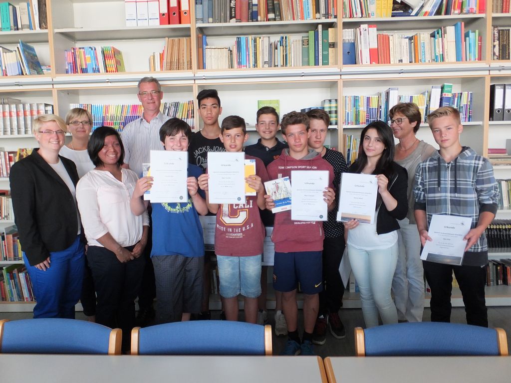 Eine Gruppe Jugendlicher hält Urkunden in die Kamera, daneben mehrere Lehrkräfte; im Hintergrund gefüllte Bücherregale der Schulbücherei.