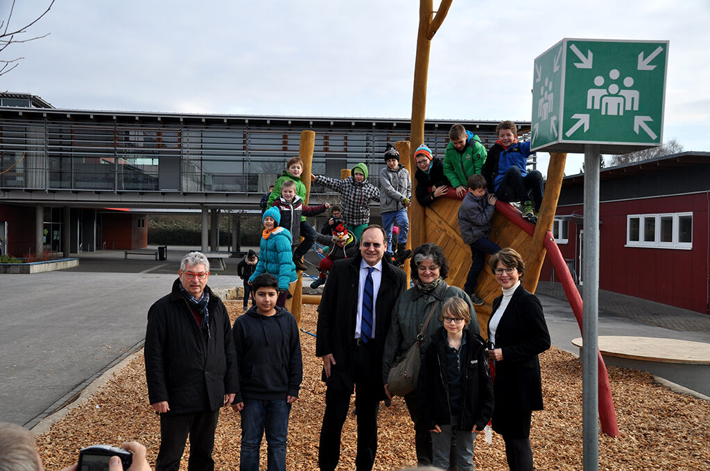 Kinder klettern auf einem hölzernen Spielgerüst, davor stehen mehrere Erwachsene; im Hintergrund das Schulgebäude und ein Sammelplatz-Schild.