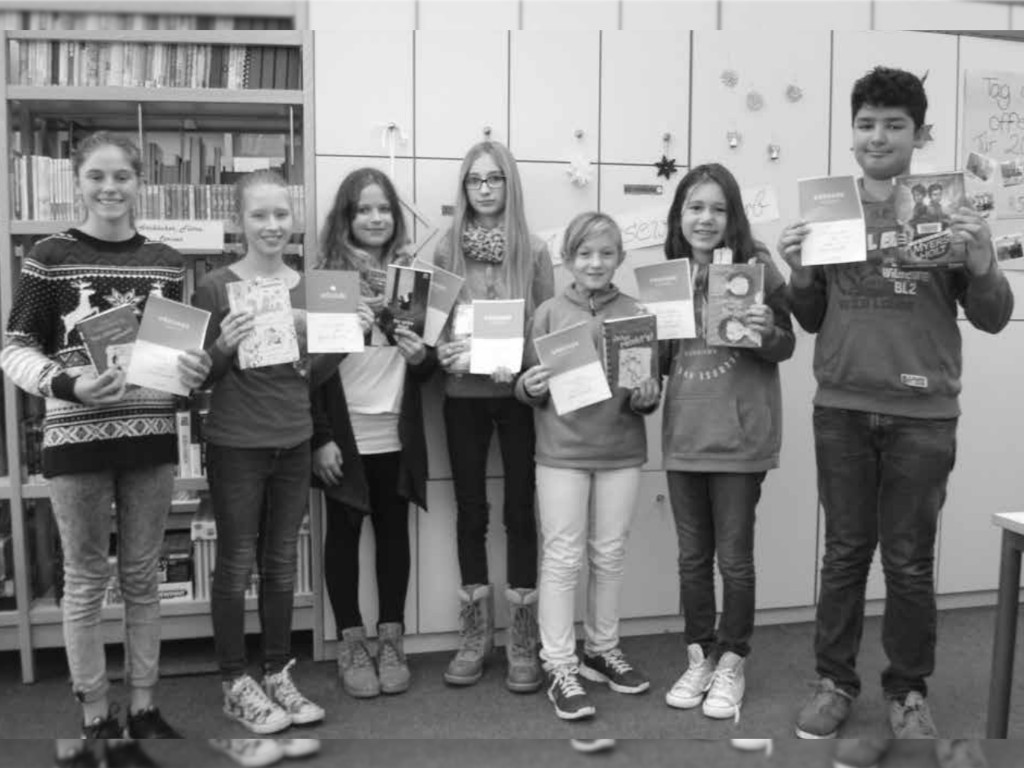 Sieben Kinder halten Urkunden und Bücher in die Kamera; im Hintergrund Bücherregale und Schränke der Schulbücherei (Schwarz-Weiß-Aufnahme).