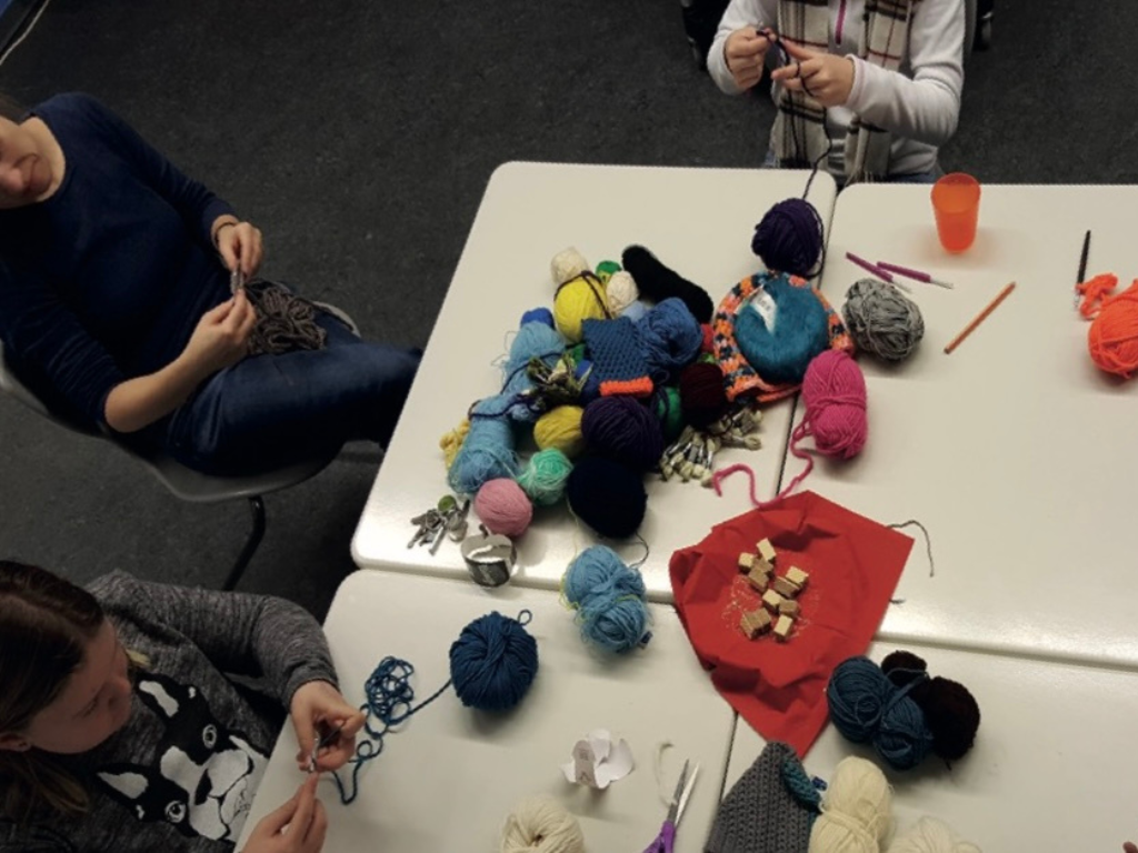 Auf einem Tisch liegen viele bunte Wollknäuel und Häkelnadeln; darum herum häkeln mehrere Jugendliche.