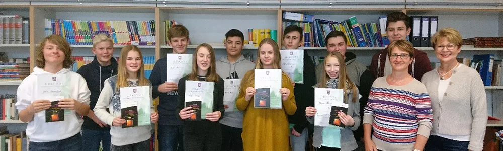 Die ausgezeichneten Schülerinnen und Schüler der 8. Klassen halten in der Bücherei ihre Urkunden und Buchpreise in die Kamera, rechts daneben zwei Lehrkräfte.