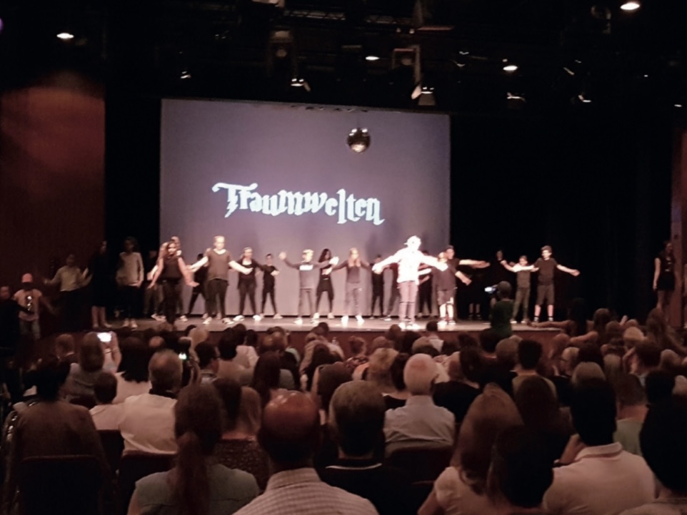 Eine Theatergruppe steht in einer Reihe mit ausgebreiteten Armen auf der Bühne vor der projizierten Aufschrift „Traumwelten“; davor ein voll besetzter Zuschauerraum.