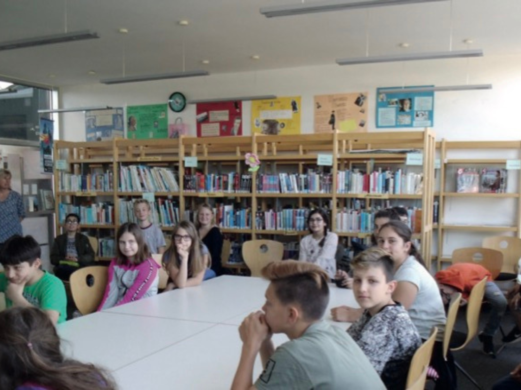 Jugendliche sitzen an Tischen in der Schulbücherei und hören einer Lesung zu; im Hintergrund gefüllte Bücherregale.
