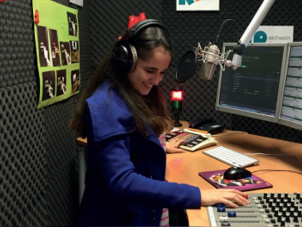 Eine jugendliche Person mit Kopfhörern sitzt an einem Mischpult mit Mikrofon und Bildschirmen im schallgedämmten Studio des Reichwein Radios.