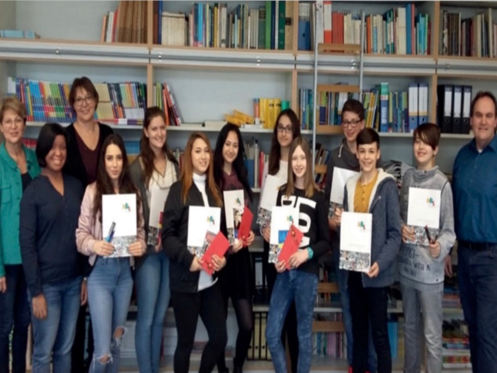Eine Gruppe Jugendlicher hält Zertifikatsmappen und kleine Geschenke in die Kamera, an den Seiten mehrere Erwachsene, vor gefüllten Bücherregalen in der Schulbücherei.