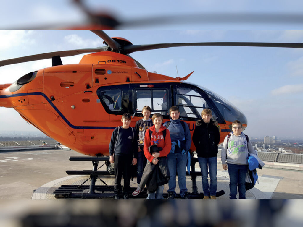 Sechs Kinder stehen vor einem orangefarbenen Rettungshubschrauber vom Typ EC 135 auf einem Dach-Hubschrauberlandeplatz; im Hintergrund eine Stadtsilhouette.