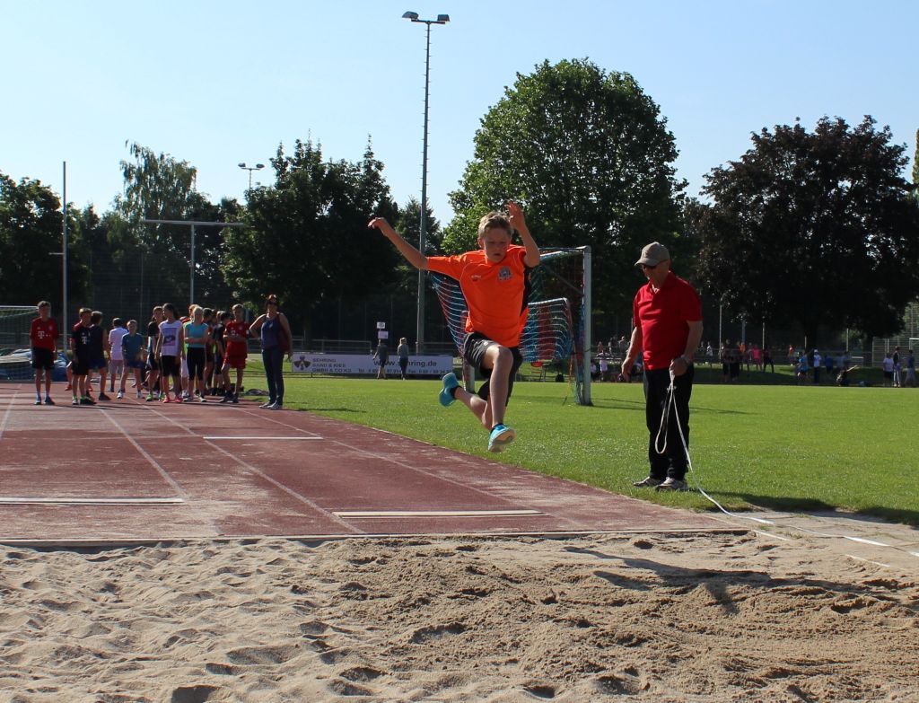 Ein Kind springt beim Weitsprung in die Sandgrube eines Sportplatzes; daneben misst eine erwachsene Person mit einem Maßband, im Hintergrund warten weitere Kinder.