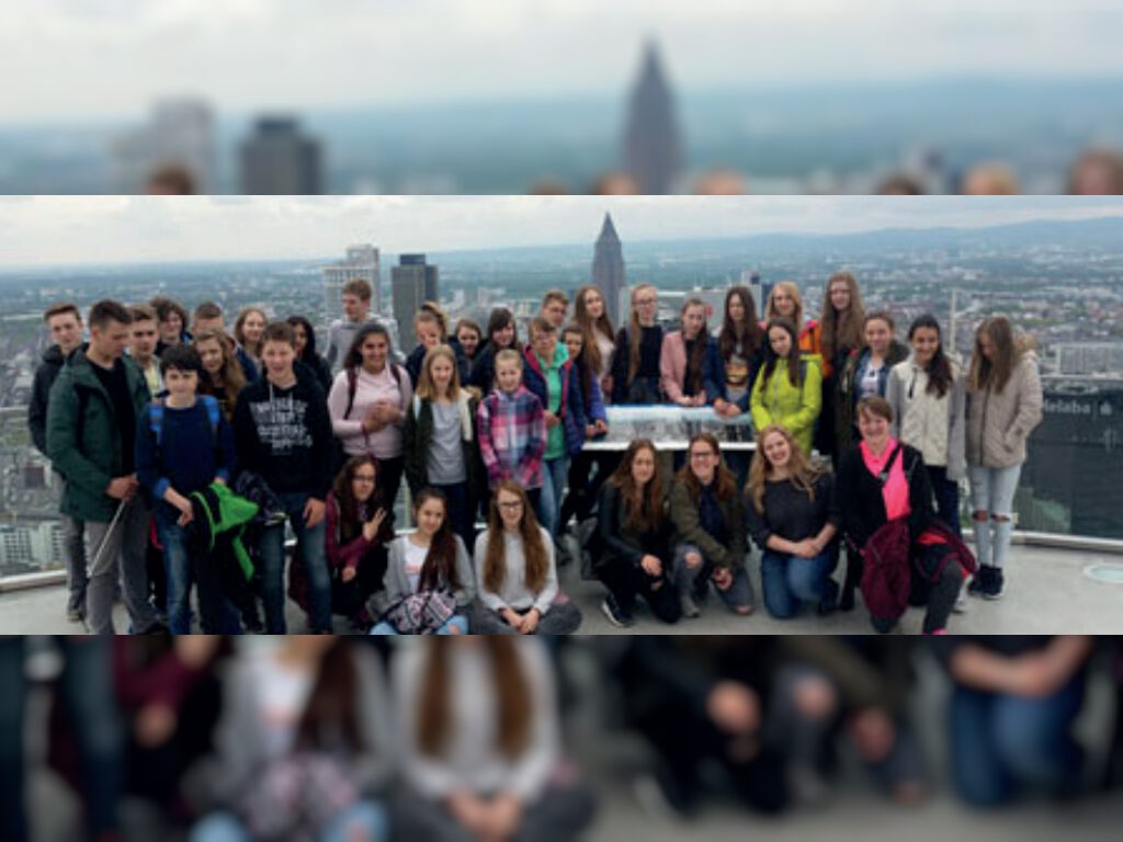 Eine große Schülergruppe posiert auf einer Aussichtsplattform hoch über Frankfurt; im Hintergrund die Skyline mit Hochhäusern und dem Messeturm.