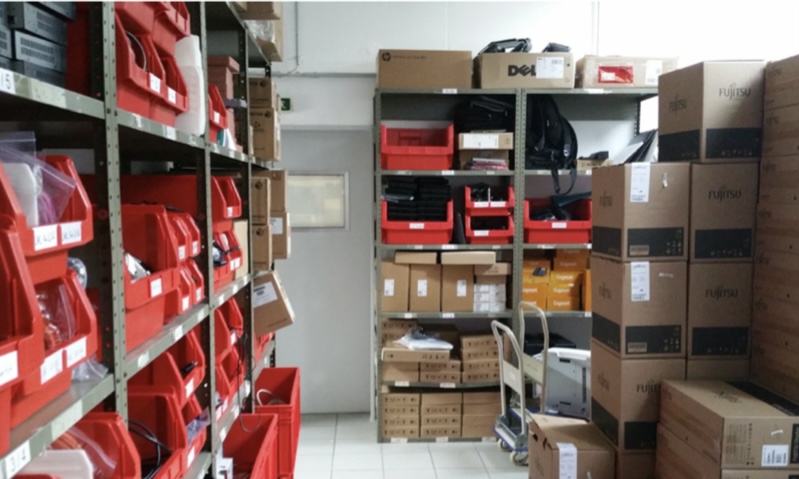Ein IT-Lagerraum mit Metallregalen voller roter Sortierboxen mit Computerteilen und Kabeln sowie gestapelten Kartons der Marken HP, Dell und Fujitsu.
