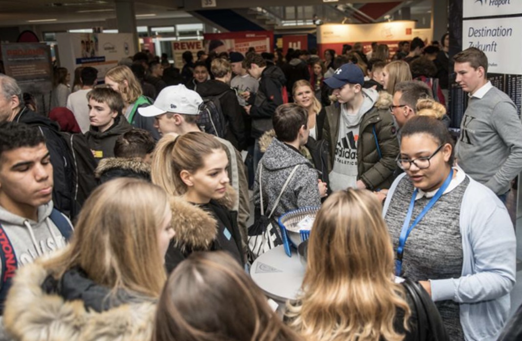 Ein dicht besuchter Ausbildungsmessestand: viele Jugendliche drängen sich zwischen Informationsständen mit Firmenlogos wie REWE und Fraport („Destination Zukunft“).