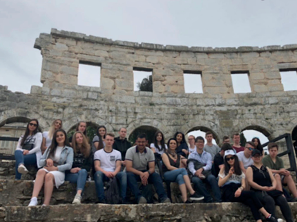 Eine Schulklasse sitzt auf den steinernen Stufen eines antiken römischen Amphitheaters; im Hintergrund die Ruinenwand der Arena.