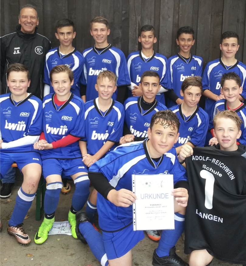 Eine Jungen-Fußballmannschaft in blauen „ARS“-Trikots posiert mit ihrem Trainer; ein Spieler hält eine Urkunde, ein anderer ein schwarzes Torwart-Trikot mit der Aufschrift „Adolf-Reichwein-Schule Langen“.