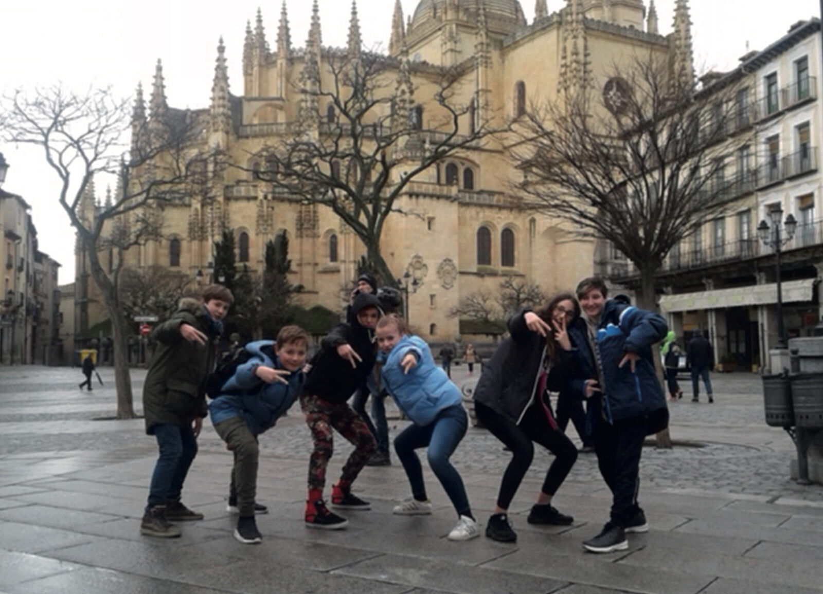 Eine Gruppe Jugendlicher posiert mit lustigen Gesten auf einem gepflasterten Platz vor einer großen gotischen Kathedrale in Spanien.