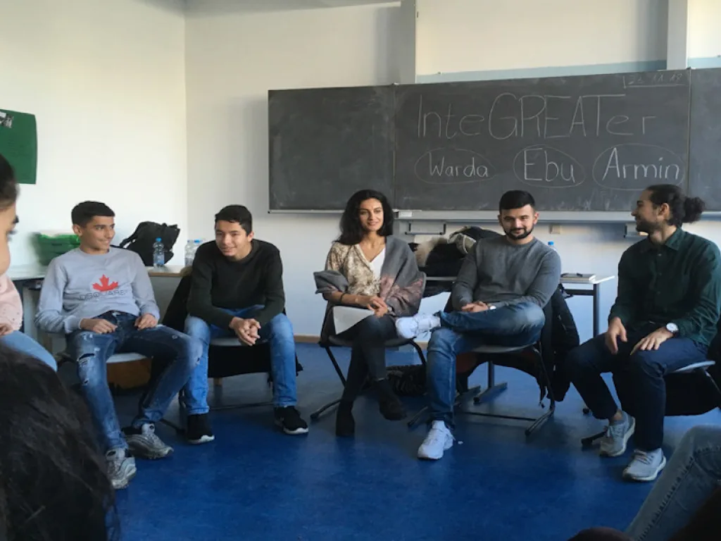 Eine Gesprächsrunde sitzt im Stuhlkreis vor einer Tafel mit dem Schriftzug „InteGREATer“.