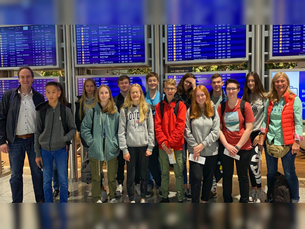 Eine Gruppe Jugendlicher posiert mit zwei Lehrkräften vor den Anzeigetafeln mit Abflugzeiten in einem Flughafen-Terminal.