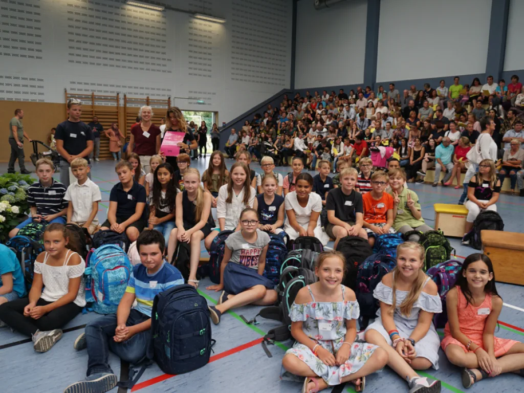 Kinder sitzen in Reihen auf dem Hallenboden, dahinter das Publikum.