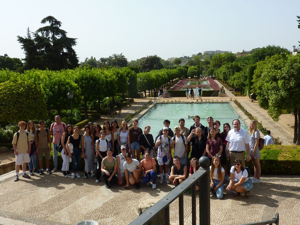 Eine große Schülergruppe posiert mit Begleitpersonen in einem spanischen Garten mit langen Wasserbecken, Springbrunnen und gepflegten Hecken.