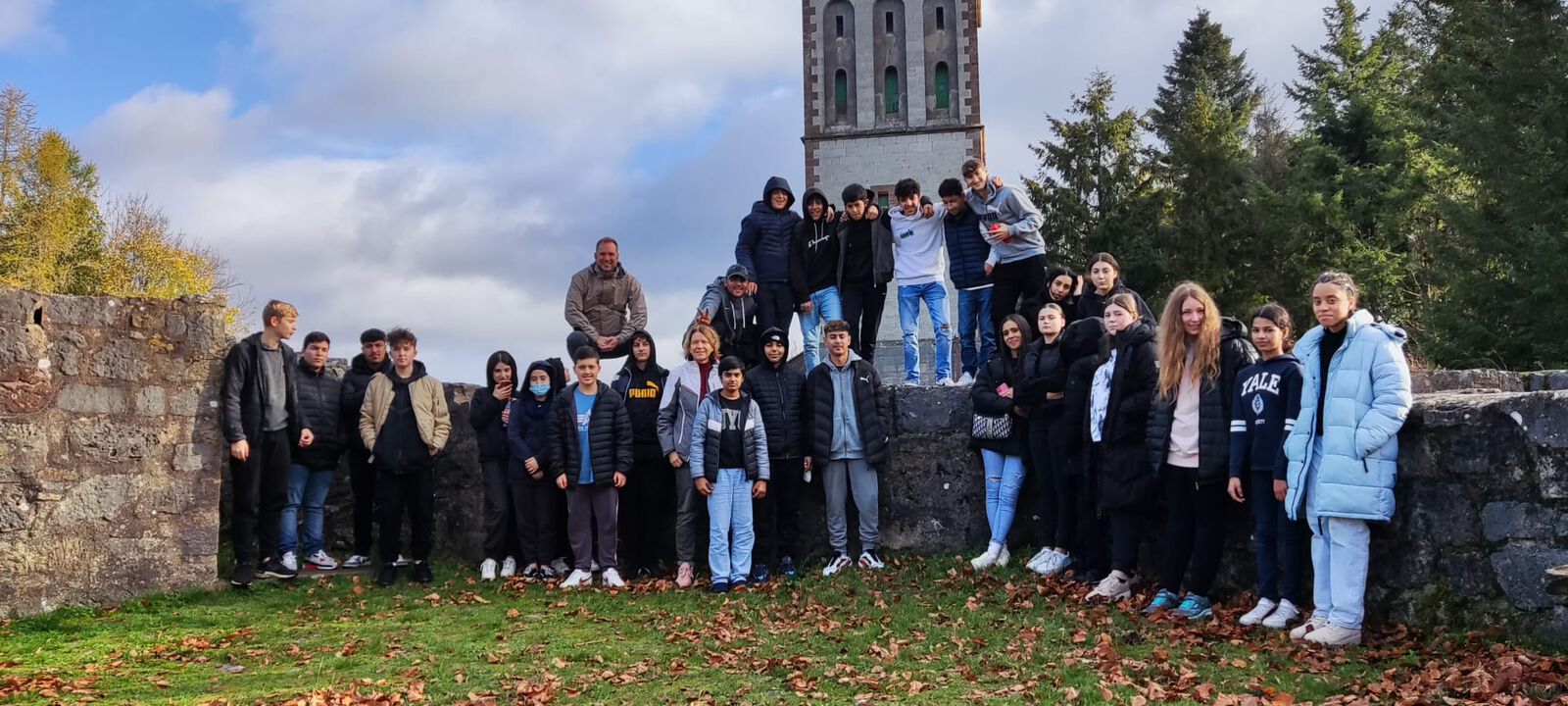 Eine Schulklasse posiert mit einer Begleitperson im Freien vor einem alten Steinturm und einer Burgruinen-Mauer; auf dem Boden liegt Herbstlaub.
