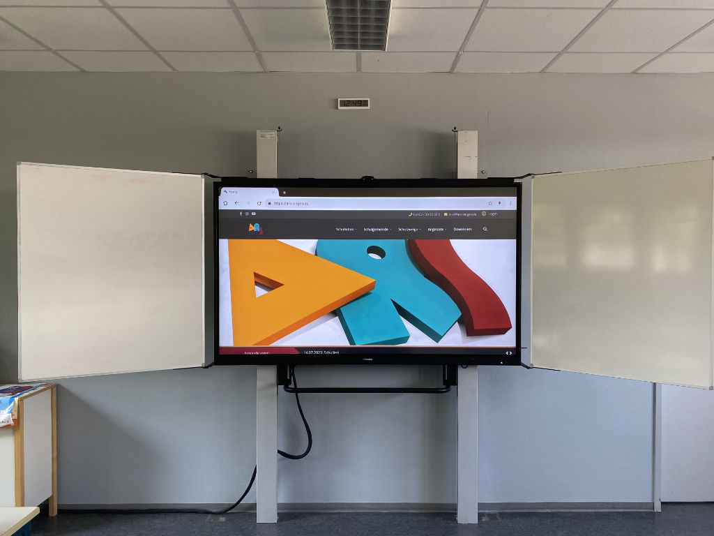 Ein großes interaktives Smartboard hängt zwischen zwei Whiteboards in einem Klassenraum und zeigt die Startseite der ARS-Website mit dem bunten Schullogo.