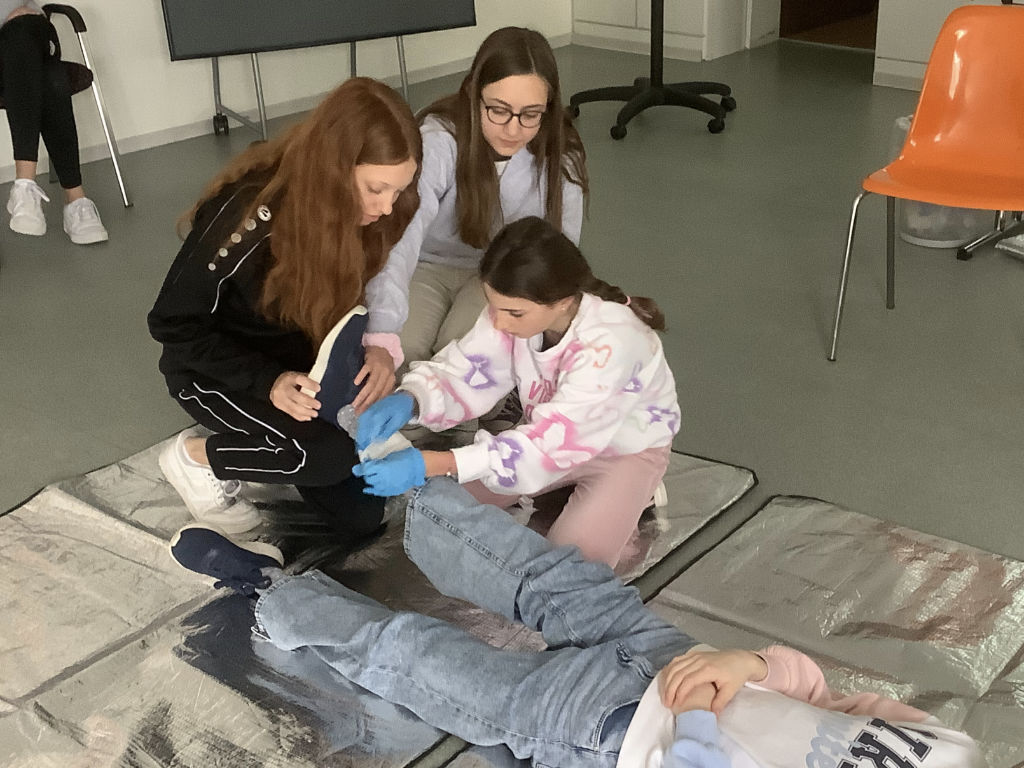Drei Jugendliche üben Erste Hilfe an einer auf einer Matte liegenden Person; eine von ihnen versorgt mit blauen Einmalhandschuhen das Knie.