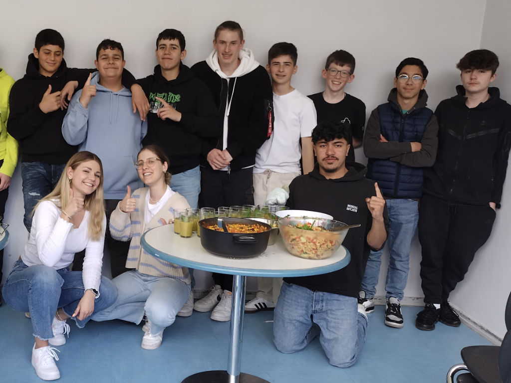 Eine Gruppe Jugendlicher posiert um einen Tisch mit selbst gekochtem Essen: ein Topf mit einem Reis-Gemüse-Gericht, eine Schüssel Salat und Gläser mit einem grünen Smoothie.