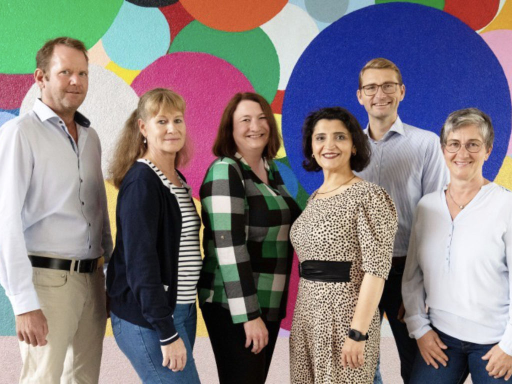 Sechs Erwachsene stehen lächelnd nebeneinander vor einer bunt mit großen Kreisen bemalten Wand.