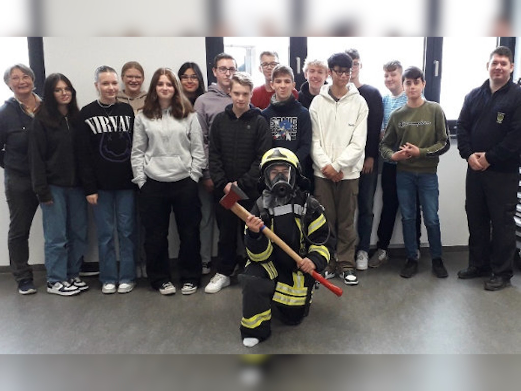 Eine Gruppe Jugendlicher steht mit zwei Erwachsenen in einem Klassenraum; vorne kniet eine Person in kompletter Feuerwehr-Schutzausrüstung mit Helm, Atemmaske und Axt.
