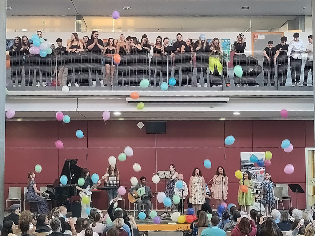 Ein Konzert im Schulforum: eine Band und mehrere Singende treten auf der Bühne auf, während zahlreiche bunte Luftballons durch die Luft schweben; von der oberen Galerie und aus dem Publikum schauen viele Menschen zu.