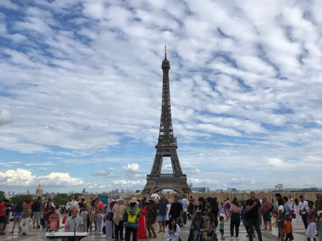 Der Eiffelturm bei Tag unter Schäfchenwolken, davor Menschen auf dem Vorplatz.