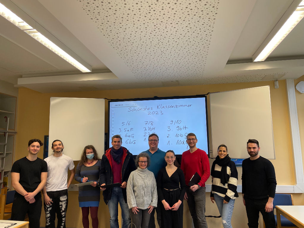 Eine Gruppe Erwachsener und Jugendlicher steht in einem Klassenraum vor einem Smartboard, auf dem die handschriftliche Auswertung „Schönstes Klassenzimmer 2023“ mit den Platzierungen zu sehen ist.